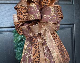May include: A decorative bow featuring a mix of leopard print and solid brown ribbon with gold accents. The bow is attached to a green wreath, creating a festive look. The bow's design includes multiple loops and streamers.