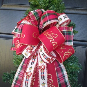 May include: A red and green plaid ribbon bow with a white ribbon featuring a repeating pattern of nutcracker figures. The red ribbon has gold glitter lettering that says "Merry Christmas".