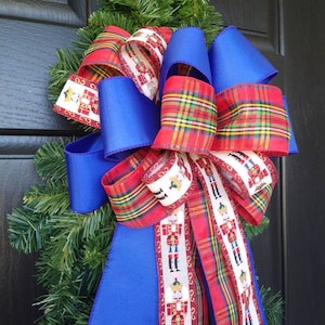 May include: A festive Christmas bow featuring vibrant blue satin ribbon and red plaid ribbon. The bow is adorned with a decorative ribbon showcasing nutcracker soldiers. The bow is attached to a green pine wreath, perfect for holiday decor.