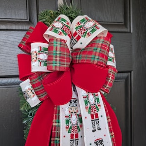 May include: A festive Christmas bow with red velvet and plaid ribbons. The bow features a cream-colored ribbon printed with nutcracker soldiers. The bow is attached to a green garland, perfect for holiday decor.