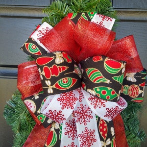May include: A red, white, and black Christmas bow with a patterned ribbon featuring ornaments and snowflakes. The bow is tied with a red sheer ribbon.
