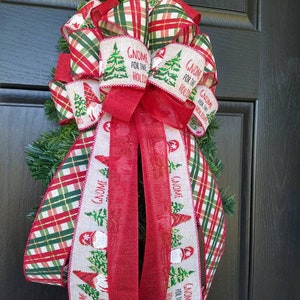 May include: A red and green plaid ribbon with a gnome design and the text "Gnome for the Holidays" is tied into a bow. The bow is attached to a green Christmas wreath.