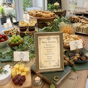 Puede incluir: Un letrero enmarcado sobre un libro dice Now Serving Breakfast, Second Breakfast, Elevenses, Lunch, Afternoon Tea, Dinner, Supper, Midnight Snack. El letrero está rodeado de varios alimentos, incluyendo uvas, queso, pastelitos y productos horneados.