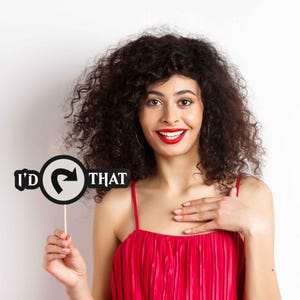 May include: A woman with curly dark hair and red lipstick smiles while holding a sign that says "I'd that". The sign is black and white with a curved arrow. She is wearing a red dress with thin straps.