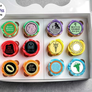 May include: A white box containing twelve cupcakes with colourful frosting and circular toppers featuring various fantasy-themed illustrations and text including Second Breakfast Approved, You Shall Not Pass Without Dessert, and One Bite To Rule Them All.