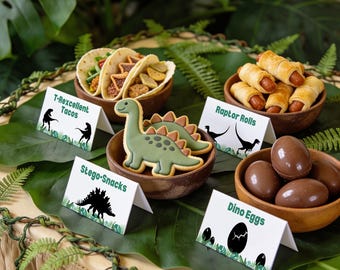 Pode incluir: Uma seleção de comida de festa com tema de dinossauros em tigelas de madeira, incluindo tacos, salsichas enroladas em massa, biscoitos verdes em forma de dinossauro e ovos de chocolate, com cartões brancos rotulados T-Rexcellent Tacos, Raptor Rolls, Stego-Snacks e Dino Eggs.