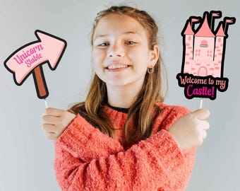 May include: A young person holds two photo booth props. One is a pink arrow sign that says "Unicorn Stable." The other is a pink castle with the text "Welcome to my Castle!" Both props are on white sticks.