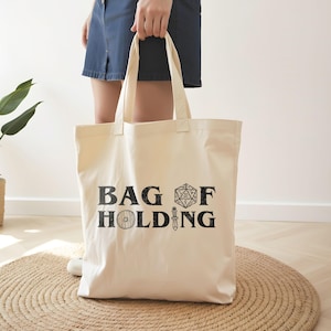 May include: A white canvas tote bag with black text that reads "Bag of Holding" and a black and white illustration of a 20-sided die and a sword.