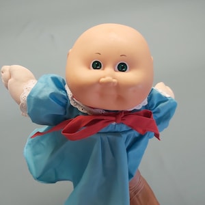 May include: A vintage Cabbage Patch Kids doll with a bald head, green eyes, and a light blue dress. The dress has a white lace collar and a red bow. The doll's arms are outstretched, and the hands are made of a soft, flesh-toned material.