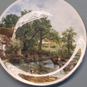 May include: A decorative plate featuring a pastoral landscape scene with a river, trees, and a small cottage. The plate has a gold rim and a detailed painting of a rural setting.