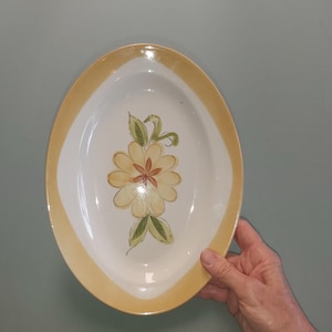 May include: Oval ceramic serving platter with a white centre and a yellow border. The platter features a hand-painted floral design in the centre with yellow petals, a red centre, and green leaves. The platter is held by a person's hand.