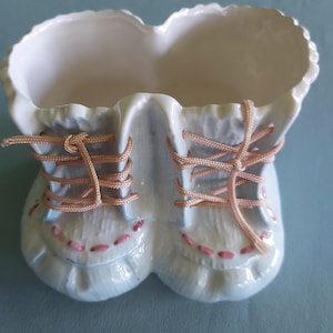 Könnte beinhalten: Ein hellblauer Keramik-Blumentopf in Form von zwei Babyschuhen. Der Pflanztopf hat rosa Nähte und pfirsichfarbene Schnürsenkel. Der obere Rand des Pflanztopfes hat ein gewelltes Design.