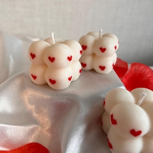 May include: Three ivory-coloured bubble candles with red heart accents. Each candle has a wick and is placed on a shiny, light-coloured surface. The candles are a decorative item, perfect for adding a touch of romance to any space.