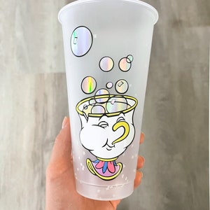 May include: A clear plastic tumbler with a white frosted finish and a design of the character Chip from Beauty and the Beast. The design features Chip with a yellow handle and a pink base, surrounded by iridescent bubbles.