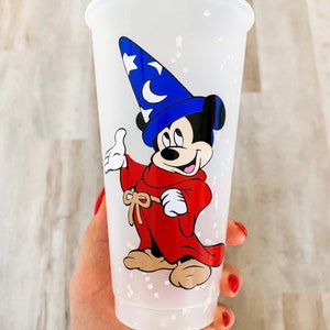 May include: A white plastic cup with a glittery finish features a cartoon image of Mickey Mouse dressed as a sorcerer. Mickey is wearing a blue hat with a star and a crescent moon, a red robe, and brown shoes.