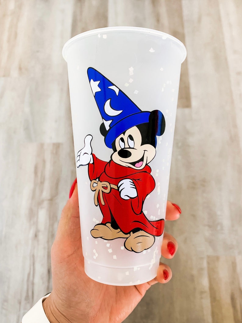 May include: A white plastic cup with a glittery finish features a cartoon image of Mickey Mouse dressed as a sorcerer. Mickey is wearing a blue hat with a star and a crescent moon, a red robe, and brown shoes.