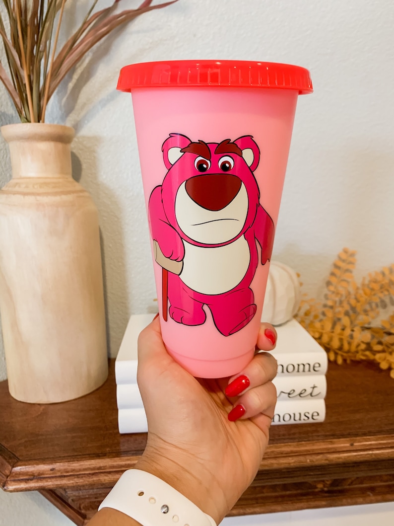 May include: Pink reusable cup with a red lid featuring a cartoon image of Lotso from the Toy Story franchise. The character is pink and has a grumpy expression.