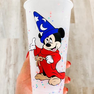 May include: A white plastic cup with a colorful confetti design. The cup features a cartoon image of Mickey Mouse dressed as a sorcerer, wearing a blue hat with a star and a crescent moon.