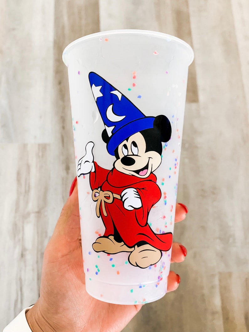 May include: A white plastic cup with a colorful confetti design. The cup features a cartoon image of Mickey Mouse dressed as a sorcerer, wearing a blue hat with a star and a crescent moon.