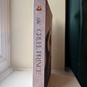 Op de afbeelding: Een hardcover boek getiteld "The Cruel Prince" van Holly Black. De rug is een gedempt roze met de titel in verticale zwarte tekst. Een eikel illustratie staat bovenaan. Het boek staat tegen een neutrale achtergrond.