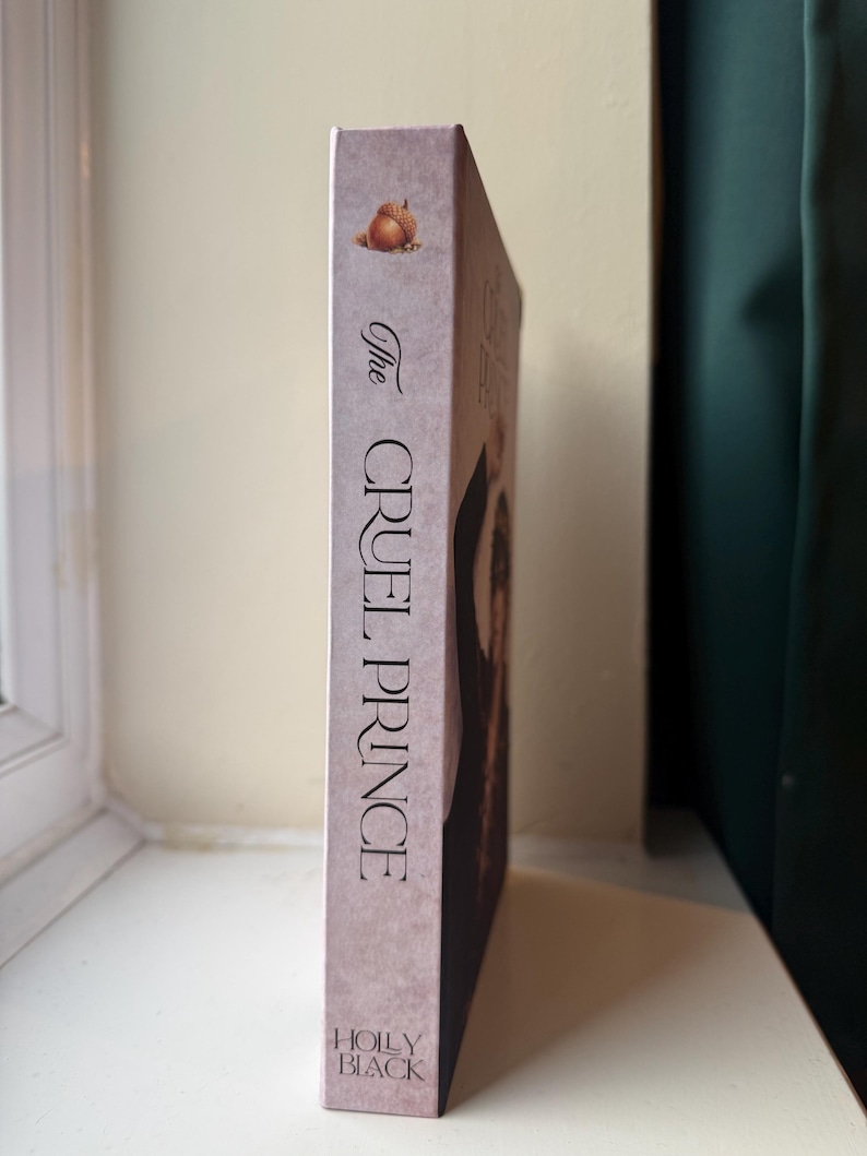 Op de afbeelding: Een hardcover boek getiteld "The Cruel Prince" van Holly Black. De rug is een gedempt roze met de titel in verticale zwarte tekst. Een eikel illustratie staat bovenaan. Het boek staat tegen een neutrale achtergrond.