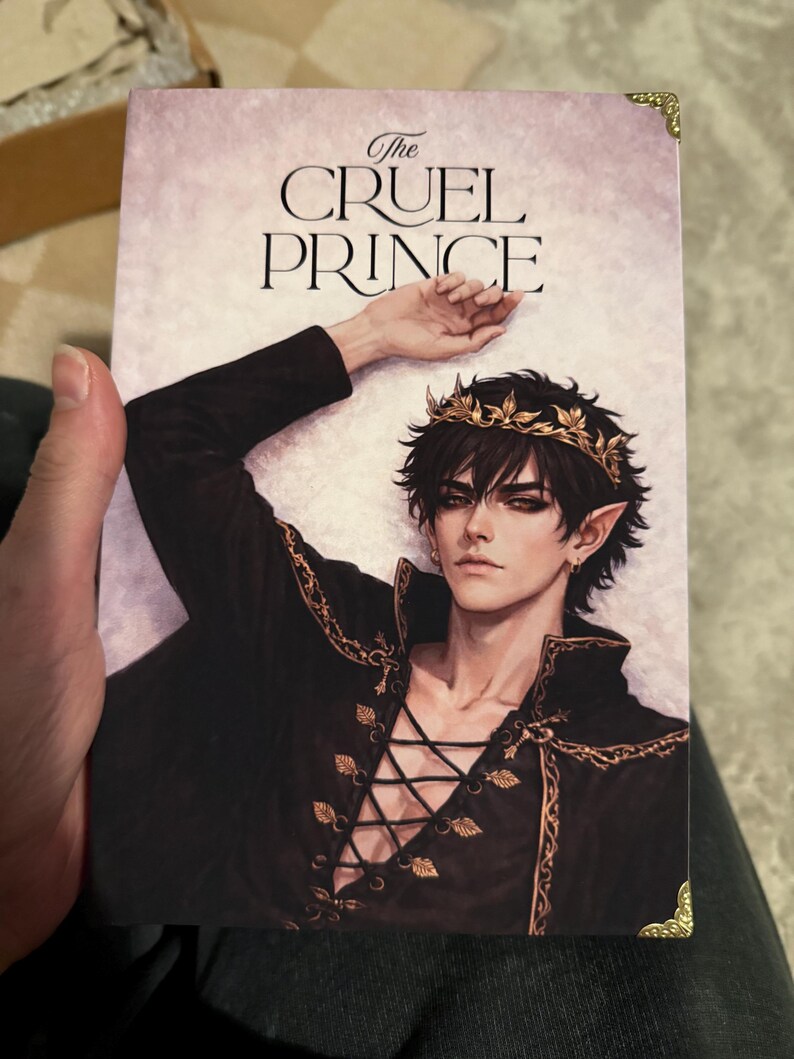 Op de afbeelding: De boekomslag van "The Cruel Prince" toont een mannelijk personage met donker haar, puntige oren en een gouden kroon. Hij draagt een zwarte outfit met gouden details en een veter aan de voorkant. De achtergrond is roze.