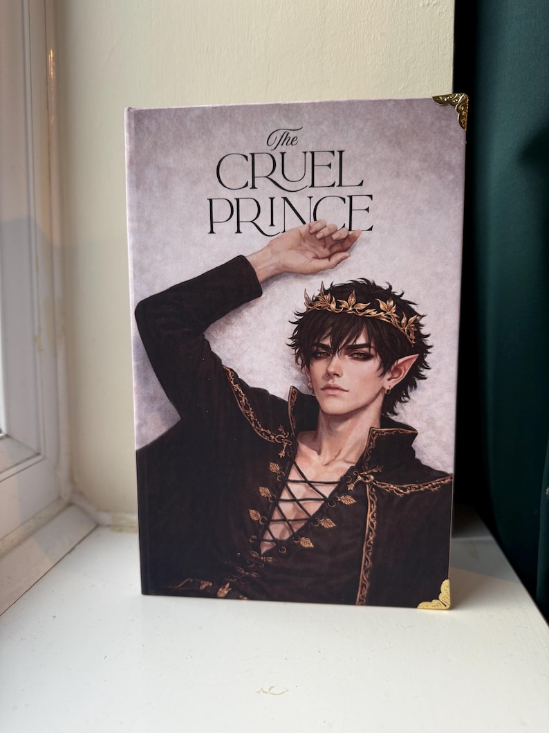 Op de afbeelding: Een hardcover boek getiteld "The Cruel Prince" met een fantasie-illustratie. De cover toont een mannelijke figuur in een zwarte outfit met gouden accenten en een kroon. Het boek heeft gouden hoekbeschermers.