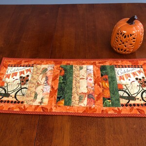 May include: A quilted table runner with a fall theme. It features a patchwork design with orange, green, and brown fabrics. The runner has a scalloped edge and is decorated with pumpkins, leaves, and a bicycle with a basket. The text "Celebrate Fall" is printed on the runner.