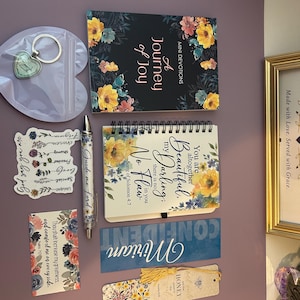 May include: A collection of stationery items including a devotional book titled A Journey of Joy, a spiral-bound notebook with floral accents, a pen inscribed with Guide me Lord, a heart-shaped keychain, a sticker, a bookmark, and a floral card.