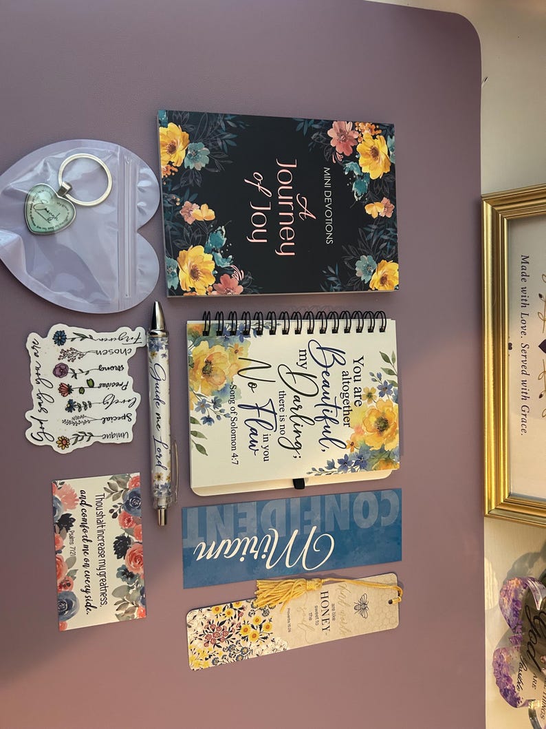 May include: A collection of stationery items including a devotional book titled A Journey of Joy, a spiral-bound notebook with floral accents, a pen inscribed with Guide me Lord, a heart-shaped keychain, a sticker, a bookmark, and a floral card.