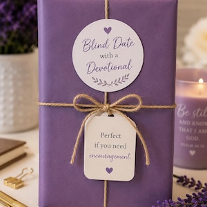 May include: A rectangular gift wrapped in solid purple paper, tied with natural jute twine. A circular white sticker reads Blind Date with a Devotional, and a rectangular white tag below reads Perfect if you need encouragement, both featuring purple text and heart icons.