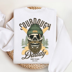 Op de afbeelding: Wit sweatshirt met een afbeelding van een schedel die een groene muts en een baard draagt. De schedel houdt twee gekruiste houten peddels vast. De tekst "Sourdough Daddy" staat in een vintage lettertype onder de schedel. De tekst "Born to Bake - Pure Instinct" staat onder de hoofdtekst.