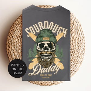 May include: Gray t-shirt with a graphic of a skull wearing a green beanie and a beard. The text "Sourdough Daddy" is printed on the shirt with the words "Born to Bake - Pure Instinct".