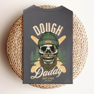 Op de afbeelding: Donkergrijs t-shirt met een afbeelding van een schedel met een groene muts en een grote snor. De tekst "DOUGH Daddy" staat boven de schedel, met "BORN TO BAKE" en "PURE INSTINCT" eronder. Twee gekruiste deegrollers staan achter de schedel.