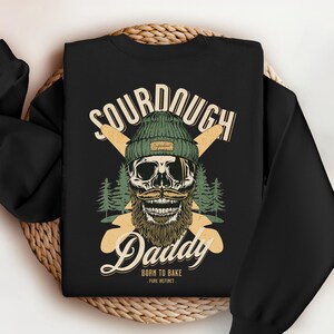 Op de afbeelding: Zwart sweatshirt met een afbeelding van een schedel met een groene en witte muts met de tekst "Sourdough Daddy Born to Bake - Pure Instinct -"