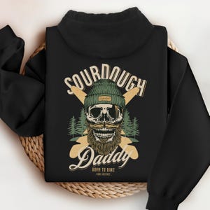 Op de afbeelding: Zwarte hoodie met een afbeelding van een schedel met een groene beanie en een snor, met de woorden "SOURDOUGH Daddy" en "Born to Bake" in vintage stijl. Twee gekruiste houten deegrollers staan achter de schedel.