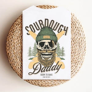 Può includere: Maglietta bianca con un design grafico di uno scheletro che indossa un berretto verde e una barba. Il testo "Sourdough Daddy" è sopra lo scheletro e "Born to Bake - Pure Instinct" è sotto lo scheletro.