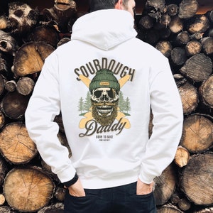 Può includere: Felpa con cappuccio bianca con una grafica di uno scheletro che indossa un berretto verde e bianco, una barba e un baffo. Il testo "Sourdough Daddy" è scritto sotto lo scheletro, con le parole "Born to Bake" e "Pure Instinct" sotto.