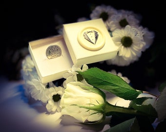 May include: A white box with a silver coin inside. The box has a small, white, raised design of a tuxedo on the lid. The box is surrounded by white flowers and a single white rose.