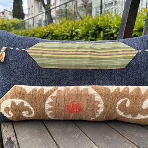 May include: A rectangular decorative pillow with a dark blue textured fabric. It features two decorative fabric patches in green and brown with a floral design. Small tassels are attached to the pillow.