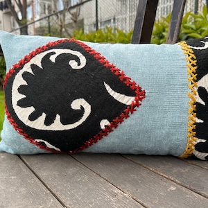 May include: A rectangular, light blue decorative pillow with a black and white patterned design. The pillow features a large, black, diamond-shaped appliqué with a white floral design and red stitching. The edges are trimmed with yellow stitching.