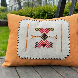 May include: An orange decorative pillow with a rectangular, off-white center panel featuring a colorful embroidered design. The design includes red, black, yellow, and orange geometric shapes. The pillow is on a wooden bench.