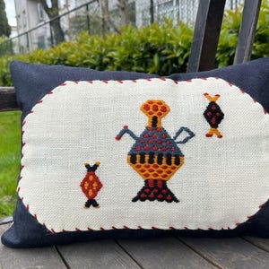 May include: A decorative pillow with a dark blue background and a white center. The center features a colorful embroidered design of a vase-like shape with arms and two fish-like shapes. The pillow is resting on a wooden bench.