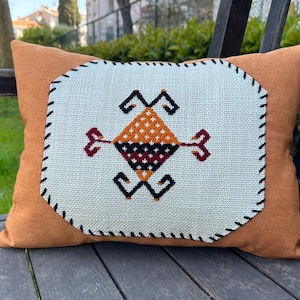 Könnte beinhalten: Ein orangefarbenes Zierkissen mit einem weißen Mittelteil, das ein geometrisches Design in Schwarz, Orange und Burgunderrot zeigt. Das rechteckige Kissen liegt auf einer Holzbank im Freien.