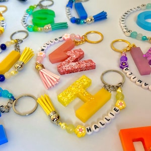 Può includere: Una collezione di portachiavi colorati con lettere e nomi. I portachiavi sono in vari colori, tra cui giallo, blu, rosa e arancione. Alcuni portachiavi hanno dettagli di perline e nappe. I nomi sono scritti con perline di lettere bianche.