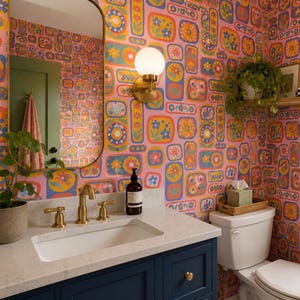 May include: A bathroom with a retro floral wallpaper in pink, yellow, blue, and orange. The room features a white sink with gold fixtures, a navy blue cabinet, and a white toilet. A mirror and a hanging plant add to the decor.