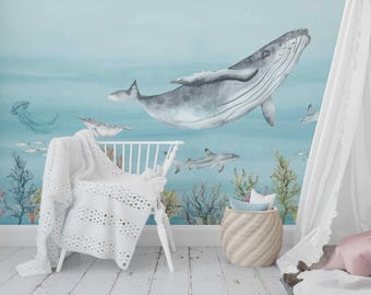 May include: A nursery scene featuring a watercolor mural of an underwater world with a whale, sharks, and other sea creatures. A white crib with a white crocheted blanket is in front of the mural. A woven basket and a tent are also in the scene.