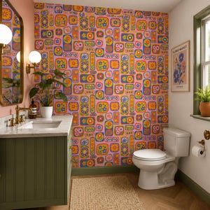 May include: A bathroom with a floral wallpaper in shades of pink, orange, yellow, and blue. The room features a green vanity with a marble countertop, a white toilet, and a framed poster. A gold-framed mirror and sconces are mounted on the pink wall.