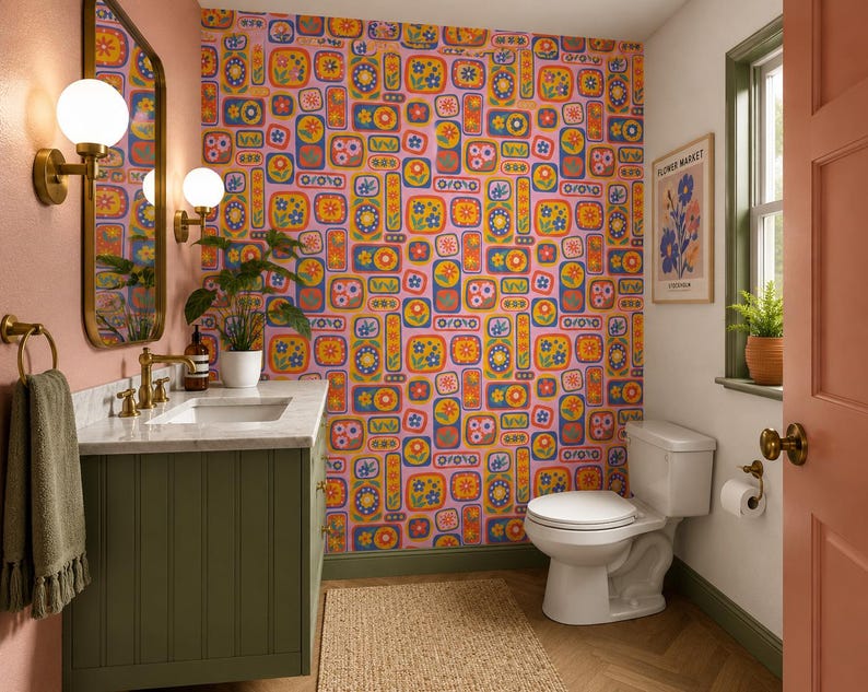 May include: A bathroom with a floral wallpaper in shades of pink, orange, yellow, and blue. The room features a green vanity with a marble countertop, a white toilet, and a framed poster. A gold-framed mirror and sconces are mounted on the pink wall.