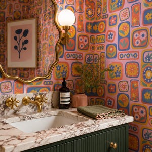 May include: A bathroom with a floral wallpaper in pink, orange, yellow, and blue. A gold-framed mirror hangs above a marble countertop with a white sink and gold fixtures. A bottle of soap and a green washcloth are on the counter.